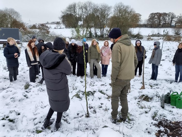 Eine Gruppe von Schülern steht im Schnee im Garten und pflanzt einen Friedensbaum.