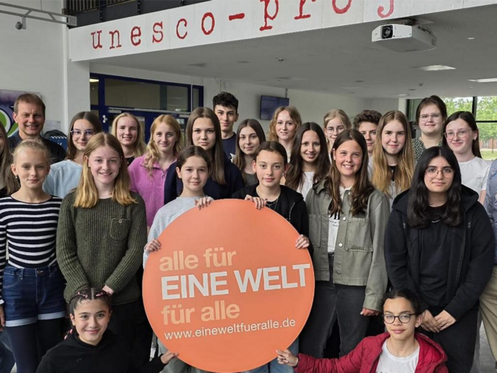 Schüler und Schülerinnen des Max-Windmüller-Gymnasium halten in dem Eingang der Schule ein Schild hoch auf dem "alle für eine Welt für alle, www.eineweltfüralle.de" steht.