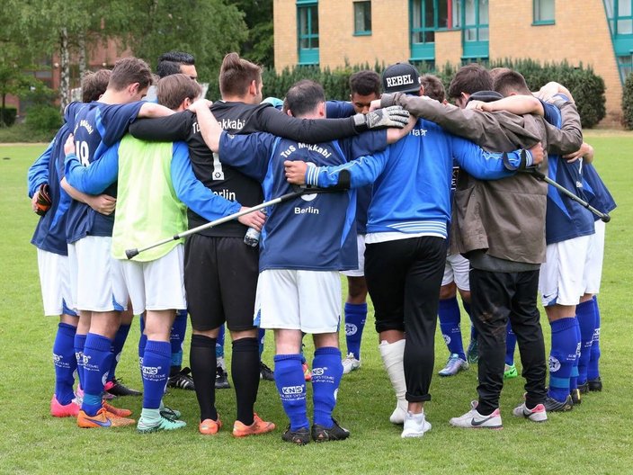 Jugendliche stehen in Fußballklamotten im Kreis auf einem Spielfeld mit Rasen. Auf den Trikots steht "Tus Maccabi Berlin".