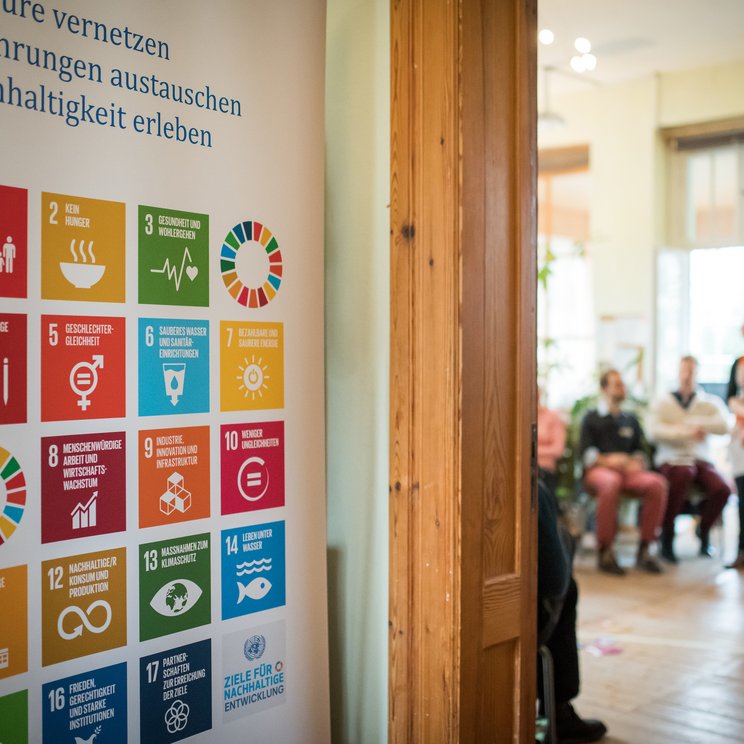 Links sieht man ein Plakat mit den SDGs und geradeaus sieht man durch einen offenen Türspalt miteinander redende Personen im Rahmen einer Tagung.