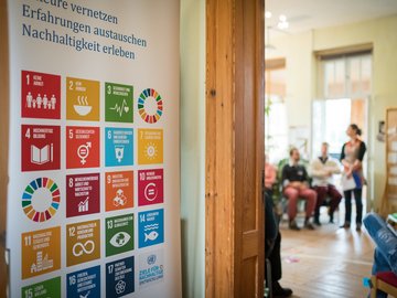 Links sieht man ein Plakat mit den SDGs und geradeaus sieht man durch einen offenen Türspalt miteinander redende Personen im Rahmen einer Tagung.