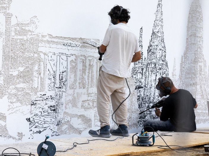Zwei Künstler arbeiten an einem Wandbild. Sie fräsen mit Bohrmaschinen die Silhouetten von Gebäuden in eine Wand.