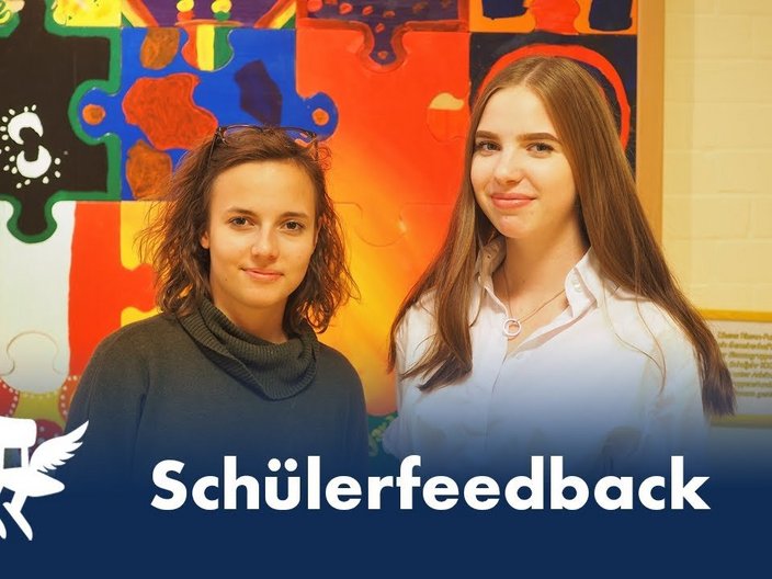 Zwei Schülerinnen stehen vor einer künstlerisch-bunt gestalteten Wand und vor ihnen steht der Schriftzug "Schülerfeedback" und links daneben ist das Zeichen des Deutschen Schulpreises abgebildet.