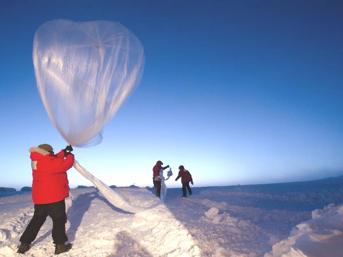 Ein Mann im Polarausrüstung steht mit dem Rücken zur Kamera und lässt einen Wetterballon in einer winterlichen Landschaft bei blauem Himmel aufsteigen.