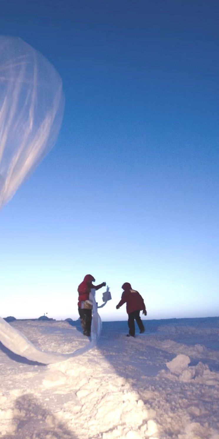 Ein Mann im Polarausrüstung steht mit dem Rücken zur Kamera und lässt einen Wetterballon in einer winterlichen Landschaft bei blauem Himmel aufsteigen.