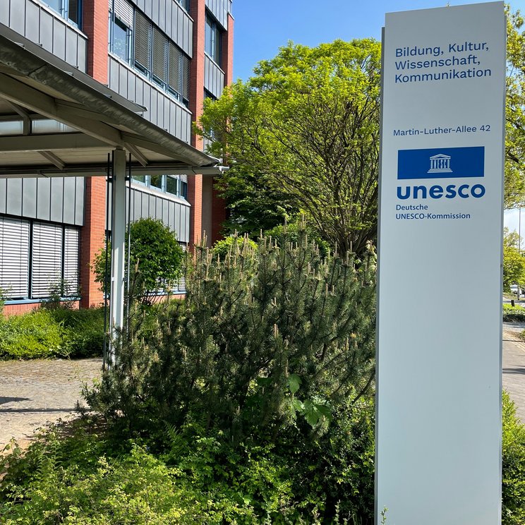 Weiße Stele mit blauem UNESCO-Logo und Adresse vor dem DUK-Gebäude