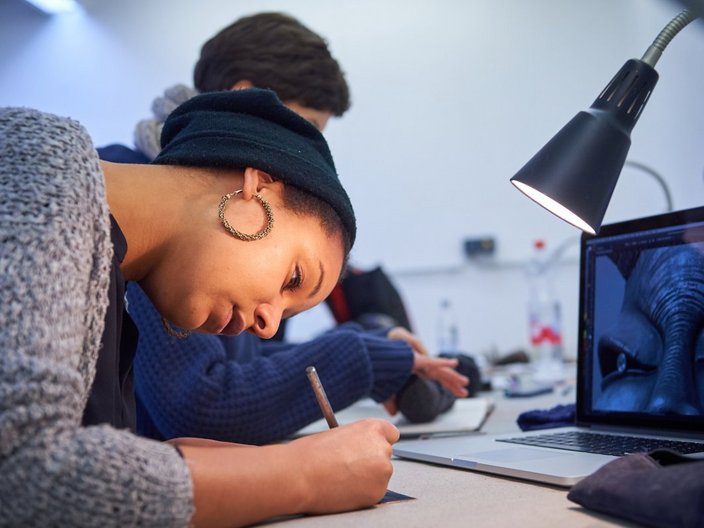 Eine Frau sitzt vor einem Laptop und schreibt in ein Heft.