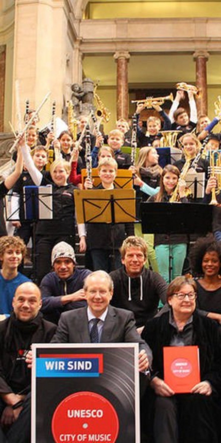 Eine Gruppe Menschen steht auf der Rathaustreppe in Hannover mit Schildern mit der Aufschrift "Wir sind UNESCO City of Music" 