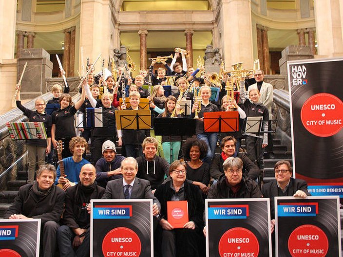 Eine Gruppe Menschen steht auf der Rathaustreppe in Hannover mit Schildern mit der Aufschrift "Wir sind UNESCO City of Music" 