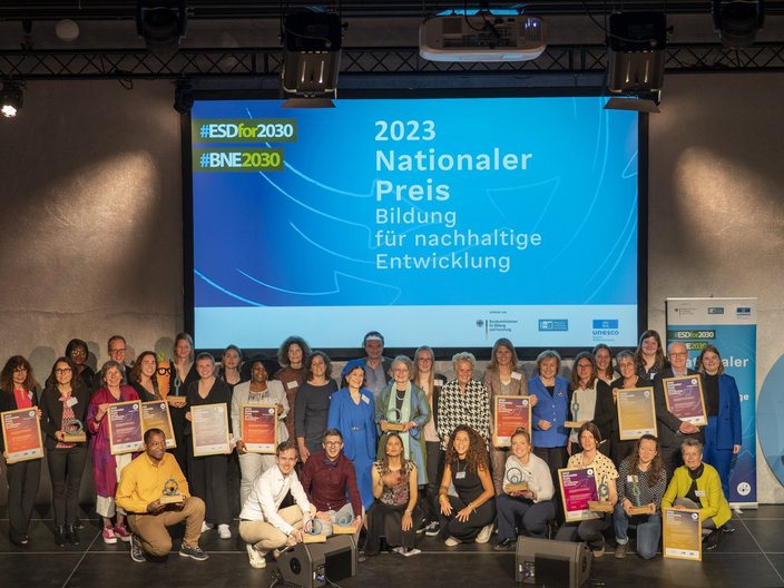 Auf diesem Foto sind die BNE-Preistragenden des Jahres 2023 mit ihren Urkunden und Trophäen auf der Bühne der Kulutrbrauerei in Berlin abgebildet