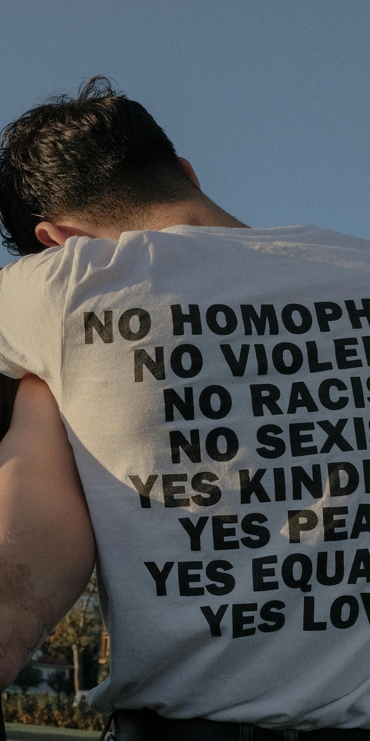 Zwei Männer lehnen aneinander. Auf dem T-Shirt-Rücken des einen Mannes steht "No homophobia, no violence, no racism, no sexism, yes kindness, yes peace, yes equality, yes love", 