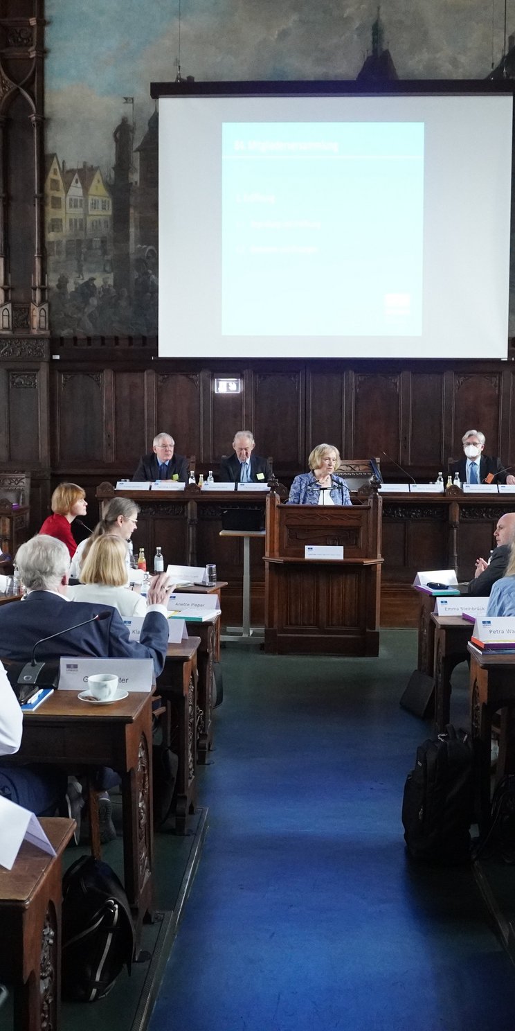 Blick in einen Plenarsaal. Im Hintergrund das Podium, an dem eine Frau zu den versammelten Menschen spricht.