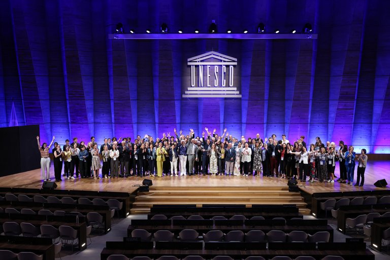 Eine große Gruppe von Menschen posiert im Plenarsaal der UNESCO auf der Bühne. Im Hintergrund ist an der Wand das Logo der UNESCO angebracht.