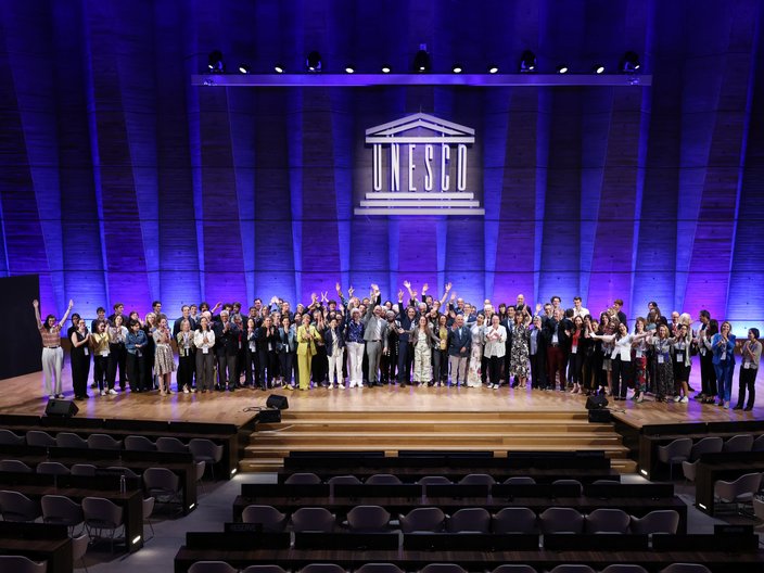 Eine große Gruppe von Menschen posiert im Plenarsaal der UNESCO auf der Bühne. Im Hintergrund ist an der Wand das Logo der UNESCO angebracht.