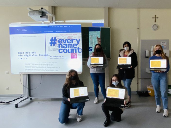 Fünf Schüler und Schülerinnen stehen vor der Tafel im Klassenzimmer und halten ihre Laptops hoch. Auf den Bildschirmen sieht man fünf gelbe Akten. Hinter den Schüler und Schülerinnen steht projiziert "#everynamecounts".