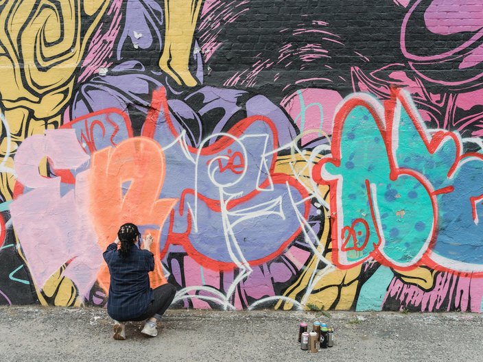 Eine junge Frau kniet vor einer bunt bemalten Wand und malt ein Graffiti darauf.