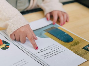 Man sieht ein Kinderbuch mit Text, Brailleschrift und gezeichneten Bildern. Ein Finger fährt über die Buchseiten. An dem Finger sieht man abgeblätterten Nagellack. Die Hand sieht aus, als gehöre sie zu einem Mädchen im Grundschulalter.