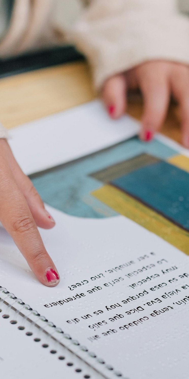 Man sieht ein Kinderbuch mit Text, Brailleschrift und gezeichneten Bildern. Ein Finger fährt über die Buchseiten. An dem Finger sieht man abgeblätterten Nagellack. Die Hand sieht aus, als gehöre sie zu einem Mädchen im Grundschulalter.