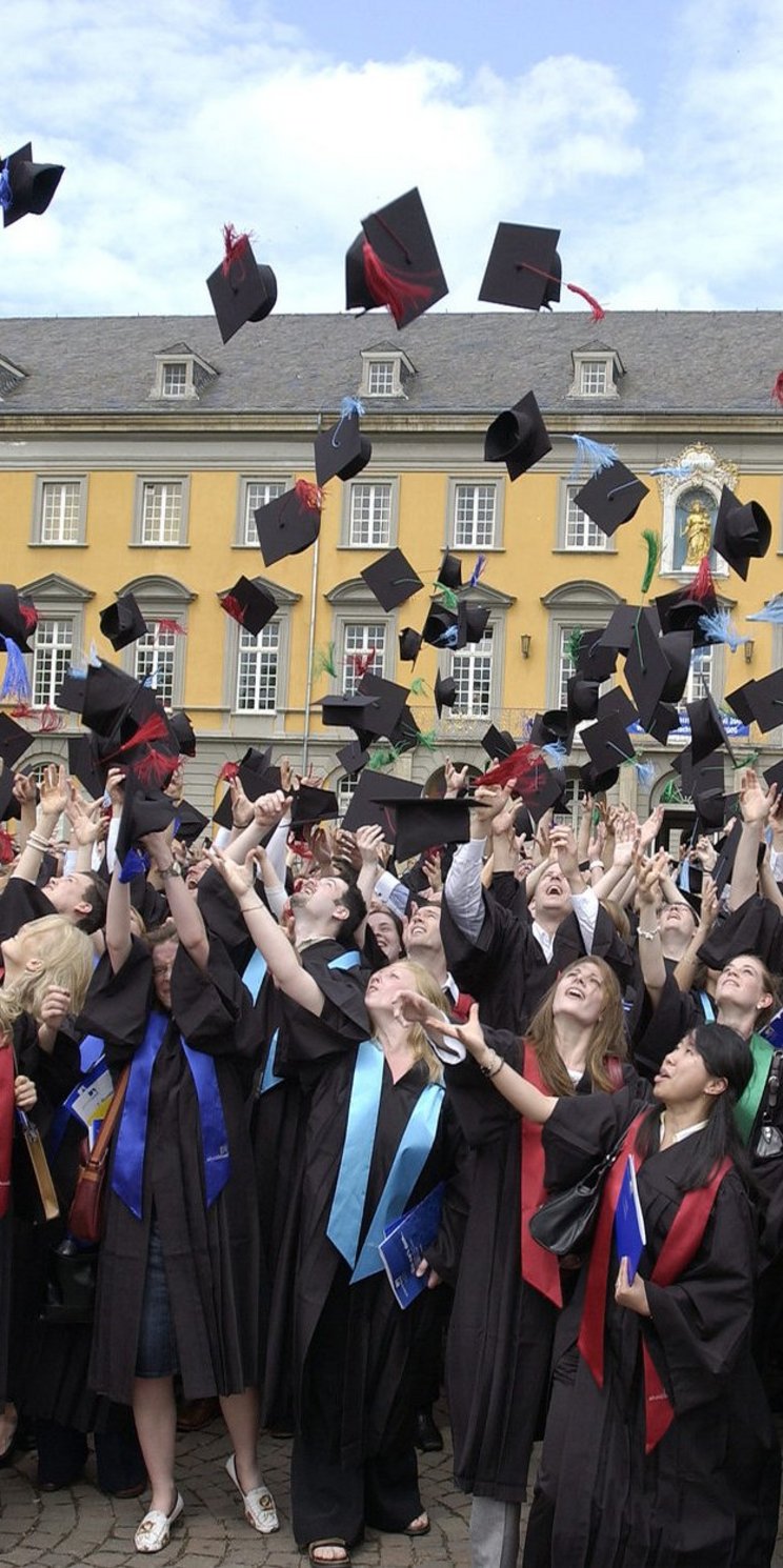 Absolventinnen und Absolventen werfen ihre Hüte vor der Universität Bonn in die Luft.