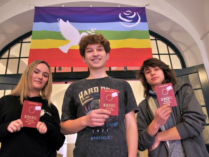 Drei Schüler*innen stehen vor einer Rainbow-Peace Flagge und halten einen Pass hoch auf dem "Krieg, stell dir vor, es wäre hier" steht.