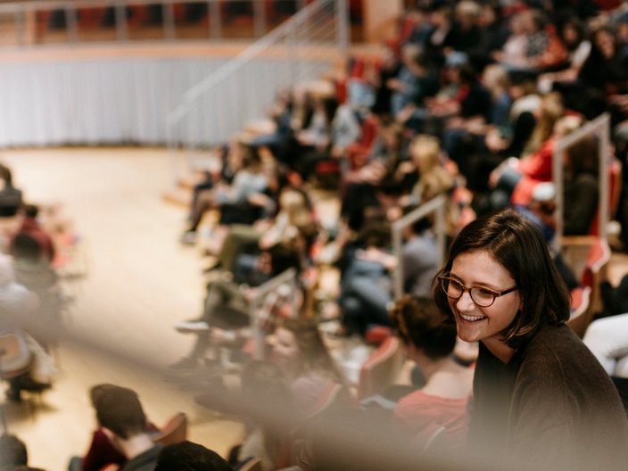 Vorlesungssaal einer Universität, die voll mit Studierenden besetzt ist. Eine Frau mit Brille ist im Vordergrund. Die Personen hinter ihr sind verschwommen. 