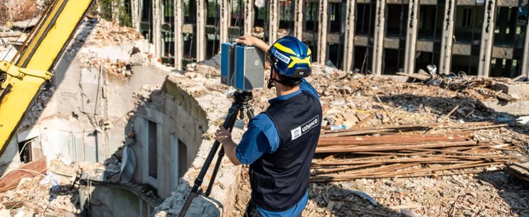 Ein Mann mit einer UNESCO-Weste und einem UNESCO-Helm steht in einer Ruine in der Ukraine und benutzt ein Vermessungsgerät.