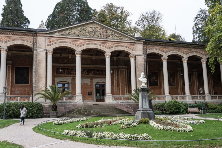 Baden-Badens Trinkhalle von außen, ein prächtiges Gebäude mit Säulengang, davor ein Garten mit einem Rondell aus Blumen. In der Mitte eine Büste des Kaisers Wilhelm I.