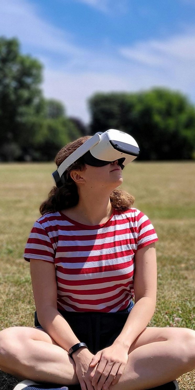 Eine Frau sitz mit einer VR-Brille auf einer Wiese vor einem Wald.