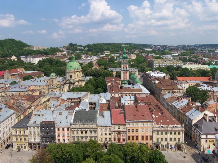 Blick von oben auf die Stadt Lwiw. Man sieht viele alte, schöne Häuser, zentral ein sakrales Kuppelgebäude und im Hintergrund grüne Hügel.