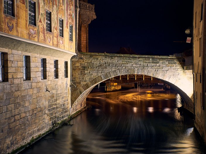 Anblick einer angestrahlten historischen Gebäudefassade mit angrenzender Brücke bei Nacht in der Altstadt von Bamberg