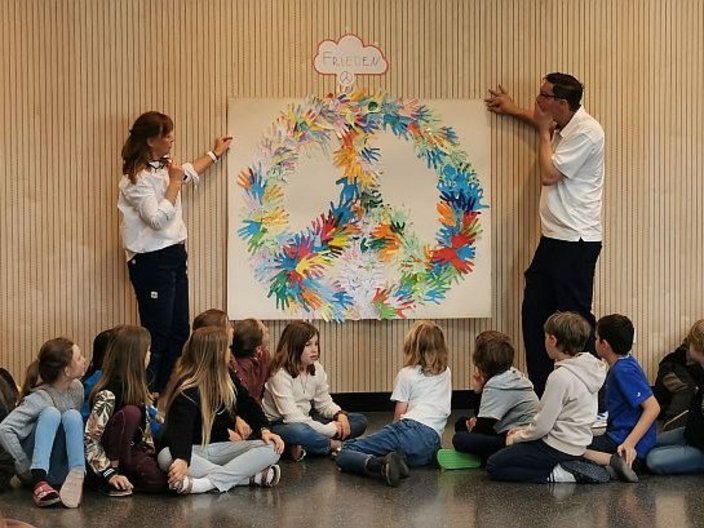 Schüler*innen schauen auf ein gemeinsam kreiertes Peace-Zeichen an der Wand welches von zwei Lehrkräften erklärt wird.