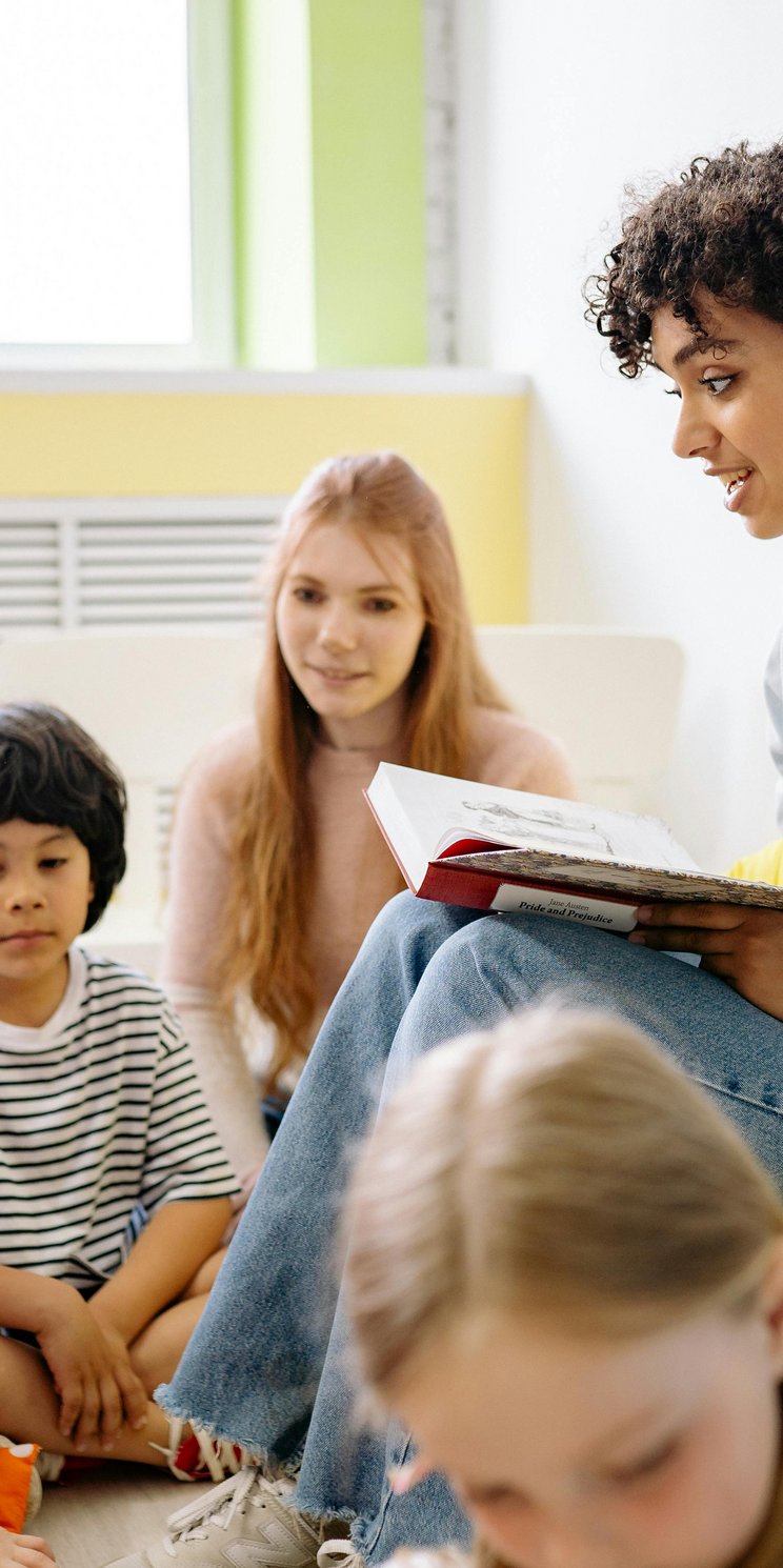 Eine Frau mit schwarzen Locken sitzt auf einem Sessel und liest mehreren Kindern im Kindergarten- oder Grundschulalter vor.