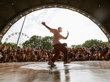 Ein junger Mann tanz auf einer Festivalbühne Breakdance. Vom Rand der Bühne schauen ihm die Zuschauer zu. 