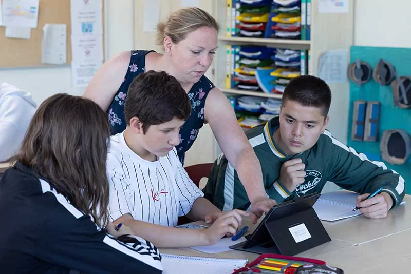 Drei Schüler sitzen an einem Tisch und die Lehrerin erklärt Ihnen etwas an einem Notebook.